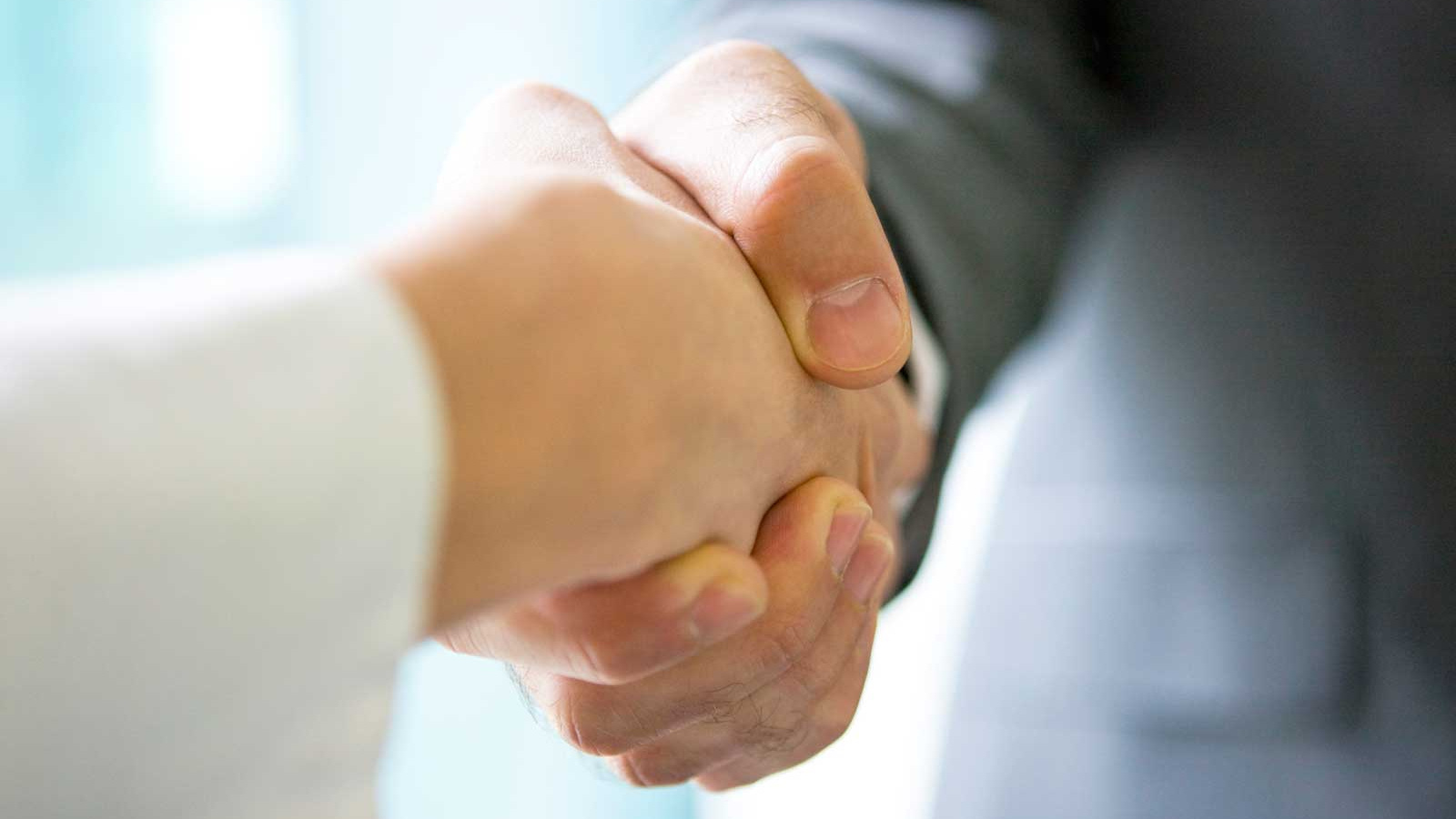 Una fotografía de primer plano de la mano de un hombre estrechando la mano de otra persona, vistiendo una camisa blanca de manga larga, en un entorno profesional.