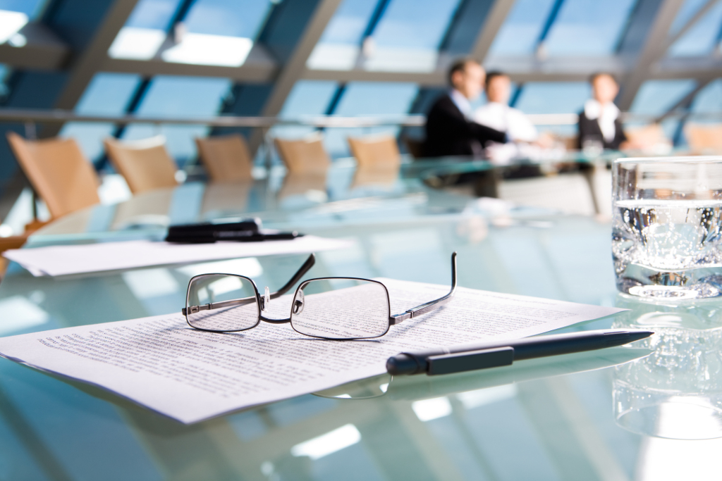 Fotografía de una moderna sala de conferencias soleada, con paredes de cristal y sillas de madera. En primer plano: una mesa de cristal con documentos, vasos y un bolígrafo. Al fondo: empresarios desenfocados, con traje, conversando.