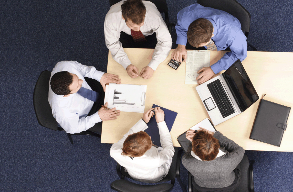 Vista aérea de un equipo de cuatro empresarios vestidos de manera formal, sentados alrededor de una mesa de madera, discutiendo documentos y computadoras portátiles.