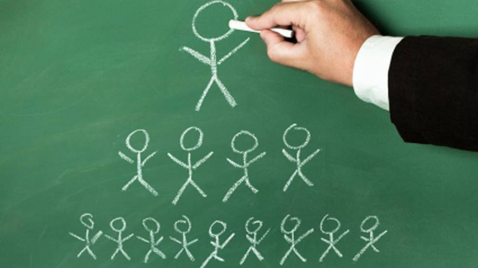 Una mano dibuja figuras de palitos en una pizarra verde, ilustrando una estructura piramidal con una figura en la parte superior y múltiples figuras debajo, que simbolizan jerarquía u organización.